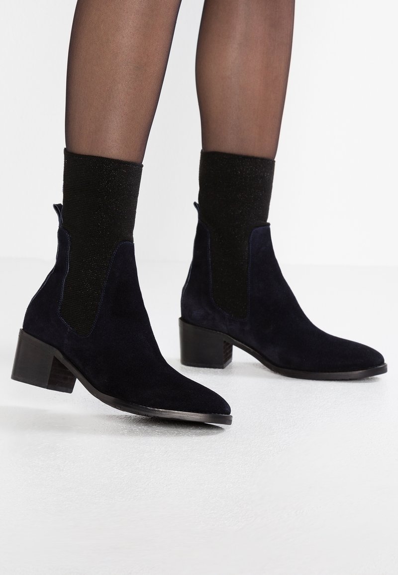 Bottes cheville en daim noir à talons blocs portées avec des collants noirs transparents et des chaussettes noires scintillantes sur fond blanc.
