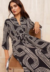 Rochie neagră cu modele ondulate crem, guler, închidere cu nasturi și detaliu legat la talie. Fabricată dintr-un material moale, cu mâneci 3/4.