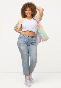 Camisa de tie-dye colorido, camiseta blanca anudada, jeans claros destrozados y zapatillas altas lilas, de pie con una mano en la cadera.