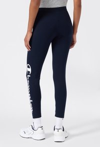Osoba ubrana w granatowe legginsy z białym logo "Champion" na lewym udzie, białe skarpetki i białe sneakersy, stojąca na jednolitym tle.