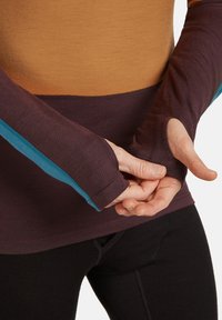 Persoon in een lange mouwen kleurblokshirt met duimgaten, die een mouw over de hand trekt, getoond van taille tot halverwege de arm.