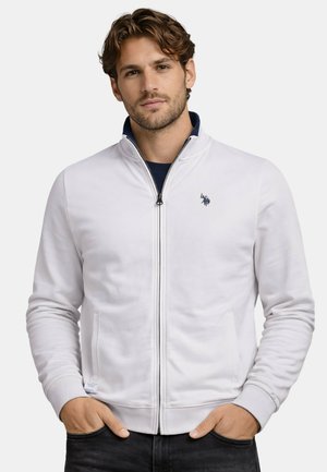 Mann trägt eine weiße Jacke mit Reißverschluss, die Hände in den Taschen, steht vor einem schlichten hellen Hintergrund.