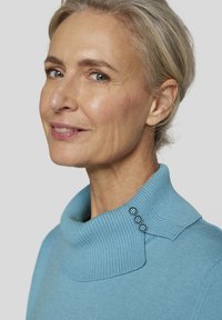 Hellblauer Pullover mit geripptem hohen Kragen und drei Knopf-Details. Weiche Textur und lockere Passform. Nahaufnahme der oberen Hälfte eines Gesichts.