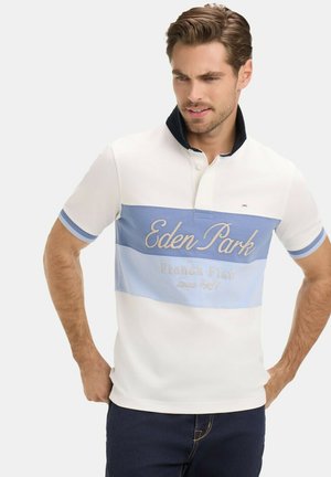 Homme aux cheveux châtain clair portant un polo blanc à rayures bleues avec le texte « Eden Park French Flair depuis 1987 », les mains sur les hanches.