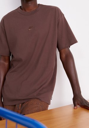 Personne portant un t-shirt Nike marron reposant une main sur une table en bois avec une poignée bleue de chariot visible au premier plan.