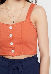 Top cropped color ruggine realizzato in denim, con spalline regolabili e bottoni bianchi sul davanti, abbinato a pantaloni corti blu navy.