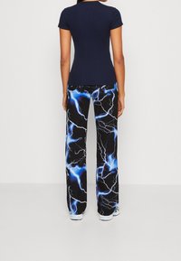 Osoba v námořnickém tričku s krátkým rukávem a černých kalhotách s modrými a bílými vzory blesků, stojící zády.