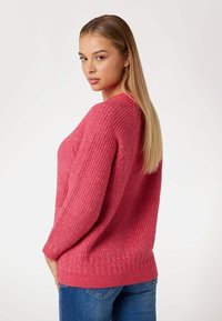 Maglione rosa lavorato a maglia con finitura testurizzata, presentante maniche lunghe e orlo a coste. Indossato sopra jeans blu.