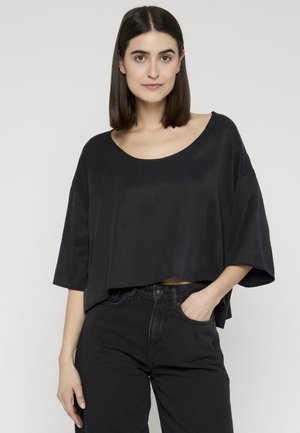 Młoda kobieta o prostych, ciemnych włosach nosi luźny, krótki czarny top i czarne jeansy z wysokim stanem, stoi z jedną ręką w kieszeni.