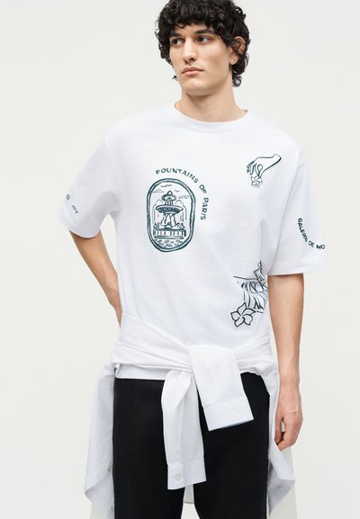 Νέος με σγουρά μαλλιά που φοράει λευκό μπλουζάκι με γραφικό σιντριβάνι "Fountains of Paris" και τυπωμένο κείμενο, με λευκό πουκάμισο δεμένο στη μέση.