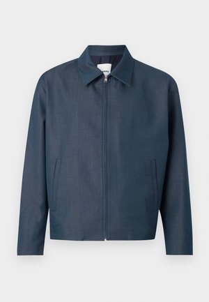 Mladý muž oblečený v tmavomodrej bunde, čierno-bielom polokošeli, bielych širokých nohaviciach a béžových topánkach stojí pred jednofarebným pozadím.