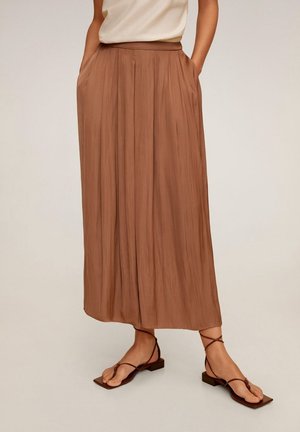 Frau trägt braune, weit geschnittene Hose mit Falten und braune flache Sandalen mit Riemchen, Hände in den Taschen, heller Hintergrund.