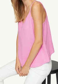 Světle růžový top bez rukávů vyrobený z texturované látky, s výstřihem do V a volnějším střihem, kombinovaný s bílými kalhotami.