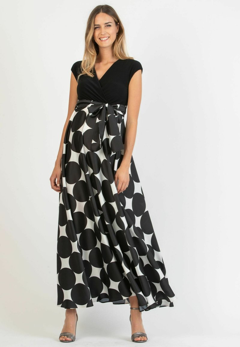 Maxi-jurk met een zwarte, getailleerde top en een vloeiende rok met grote zwart-witte cirkels. Inclusief een strik op de taille en zilveren schoenen.