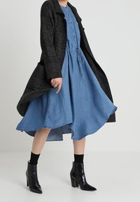 Robe en jean avec un ourlet évasé, une taille cintrée et des boutons. Superposée d'un manteau noir texturé et assortie à des bottines à talons noires.