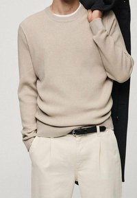 Pullover beige a coste con collo a giro, indossato sopra una canottiera bianca. Abbinato a pantaloni beige chiaro e un cappotto nero appoggiato sulla spalla.
