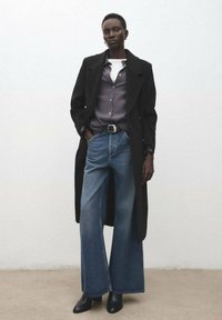 Czarny długi płaszcz, szara koszula nałożona na biały top, szerokie niebieskie dżinsy o prostych nogawkach, czarne botki i srebrna sprzączka paska. Gładkie tło.