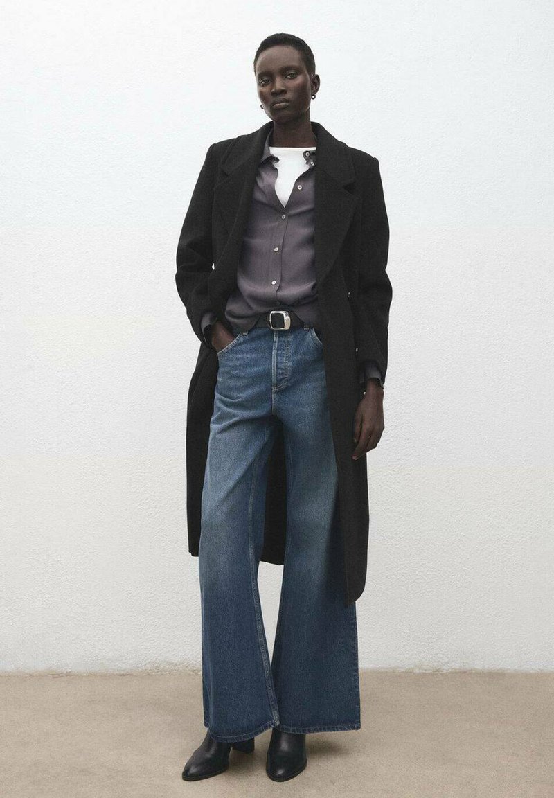 Czarny długi płaszcz, szara koszula nałożona na biały top, szerokie niebieskie dżinsy o prostych nogawkach, czarne botki i srebrna sprzączka paska. Gładkie tło.