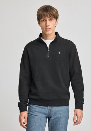 Sudadera negra con cremallera en el cuello y un logo blanco en el pecho, con puños y cintura acanalados, combinada con jeans de color azul claro.