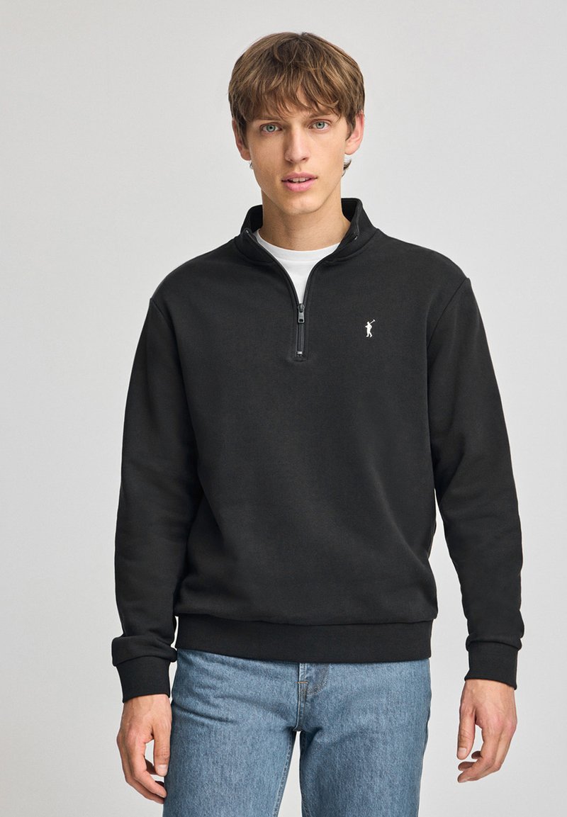 Zwarte sweatshirt met rits en een wit logo op de borst, met ribgebreide manchetten en tailleband, gecombineerd met lichtblauwe jeans.