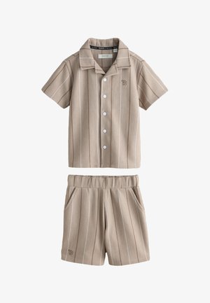 Junge läuft drinnen und trägt ein beiges kurzärmeliges Nadelstreifenhemd und passende Shorts mit weißen Socken und Turnschuhen.