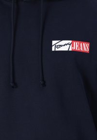 Mørkeblå hettegenser med et frontlogo med røde og hvite detaljer, snorer og et mykt, teksturert stoff.