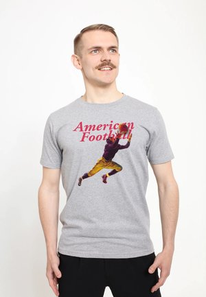 Mand med moustache iført grå T-shirt med motiv af en vintage fodboldspiller, der dykker for at gribe en bold, mod en hvid baggrund.