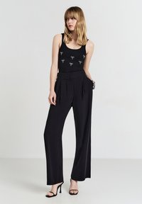 Top senza maniche nero con un motivo a fiocco decorativo, abbinato a pantaloni larghi neri a vita alta e sandali neri con cinturini.