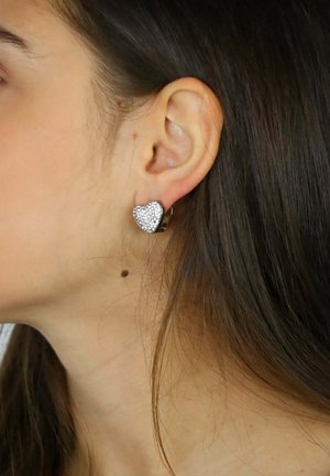Pendientes en forma de corazón en plata con piedras de cristal transparentes, con una superficie texturizada y un acento negro, mostrados en la oreja de una modelo.