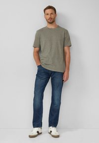 Gestreiftes graues und grünes Rundhalsausschnitt-T-Shirt kombiniert mit blauen Jeans und weißen Sneakers mit braunen Akzenten, stehend vor einem grauen Hintergrund.