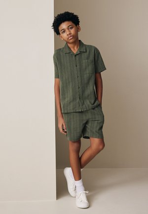 Junge lehnt an der Wand und trägt ein grünes kurzärmeliges Hemd mit Knöpfen und passende Shorts mit weißen Socken und weißen Turnschuhen.