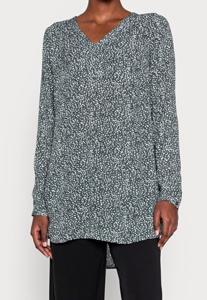 Tunique manches longues vert foncé avec motif de points blancs irréguliers, col en V, portée avec un pantalon noir, modèle debout les mains détendues.