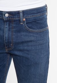 Mörkblå denimjeans med en ljus stonewash-finish. Funktioner inkluderar en framficka, kopparfärgad knapp, och synlig sömnad längs sömmarna.