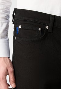 Jeans en denim noir au design ajusté. Les caractéristiques incluent une étiquette bleue, des coutures visibles et des accents en métal sur les poches.