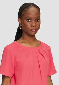 Korallenrosa Bluse mit kurzen Ärmeln, ausgestattet mit einem runden Ausschnitt und dezenten Falten an der Vorderseite. Glatte Stofftextur und lässige Passform.