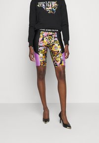 Černá mikina s nápisem "VERSACE JEANS COUTURE", barevně vzorované cyklistické šortky s černými a fialovými akcenty, doplněné černými vysokými podpatky.