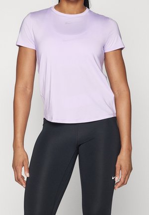 Vrouw met een lichtpaars t-shirt met korte mouwen en zwarte Nike-leggings, staand met ontspannen armen tegen een effen achtergrond.