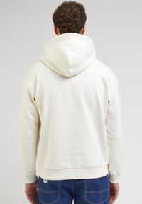 Sweat à capuche en coton crème avec une grande capuche ajustable, des poignets et un ourlet côtelés, présentant une étiquette logo discrète près de la couture de la taille.