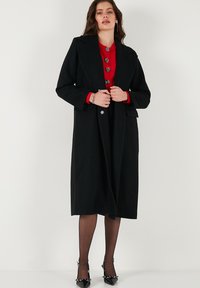 Pardesiu negru din lână, cu rever mari și o siluetă lungă, având nasturi argintii și o textură elegantă. Purtați deasupra unei bluze roșii și unei fuste negre.