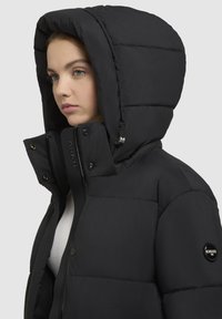 Sort pufjakke med en stor, polstret hætte. Har lynlås, trykknapper og et logo-patch på venstre ærme. Glat struktur på ydersiden.