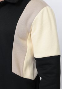 Haut à manches longues avec motif en bloc de couleurs en tissu noir, beige et crème sur le haut du bras et l'épaule.