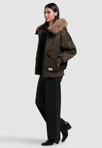 Tamsiai alyvuogių žalios spalvos bomber tipo striukė su dirbtiniu užkulniu, užtrauktuku ir priekinėmis kišenėmis. Pora su plačiaaugiu juodu kelnių ir aukštakulniais batais.