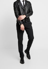 Zwarte leren motorjack met zilveren ritsen, gecombineerd met op maat gemaakte zwarte broeken en zwarte leren schoenen. Witte shirt en zwarte stropdas zichtbaar.