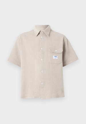 Chemise beige à manches courtes boutonnée, tissu texturé, col pointu et une poche poitrine avec un bouton, ornée d'une petite étiquette blanche et bleue.