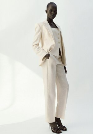 Donna che indossa un completo sartoriale color crema con blazer, gilet abbottonato, pantaloni dritti e tacchi alti neri aperti davanti, in piedi contro uno sfondo bianco.