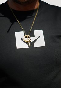 Goldene Halskette mit einem runden Anhänger, der ein erhabenes Symbol zeigt. Getragen über einem schwarzen T-Shirt mit einem weißen Grafikdesign.