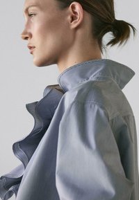 Vrouw met naar achteren getrokken haar, gekleed in een lichtblauw shirt met ruches en een hoge kraag, naar links kijkend tegen een effen achtergrond.