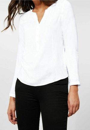 Blouse blanche à manches longues avec un décolleté en V et une patte de boutonnage, présentant un ourlet légèrement arrondi. Portée avec un pantalon noir ajusté. Texture lisse.