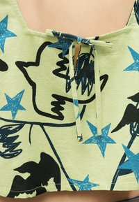 Haut en tissu vert avec des silhouettes d'oiseaux noirs et des étoiles bleues, doté d'un cordon à nouer au niveau de l'encolure.