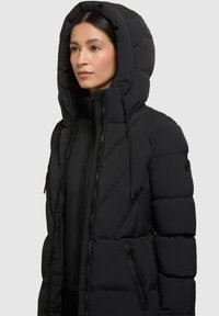 Chaqueta acolchada negra con capucha, que presenta un diseño acolchado, bolsillos con cremallera y cordones ajustables. Textura de tela suave.